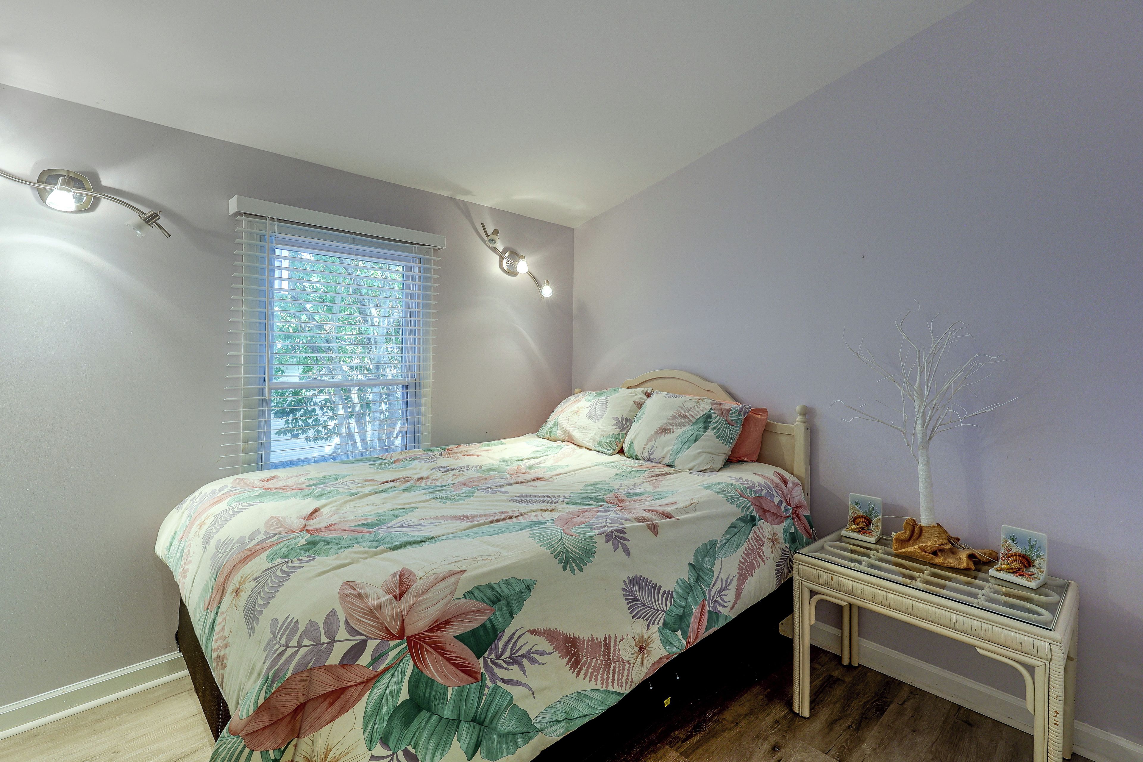 A bedroom with a bed, a nightstand, a window with blinds, and a wall-mounted light fixture.