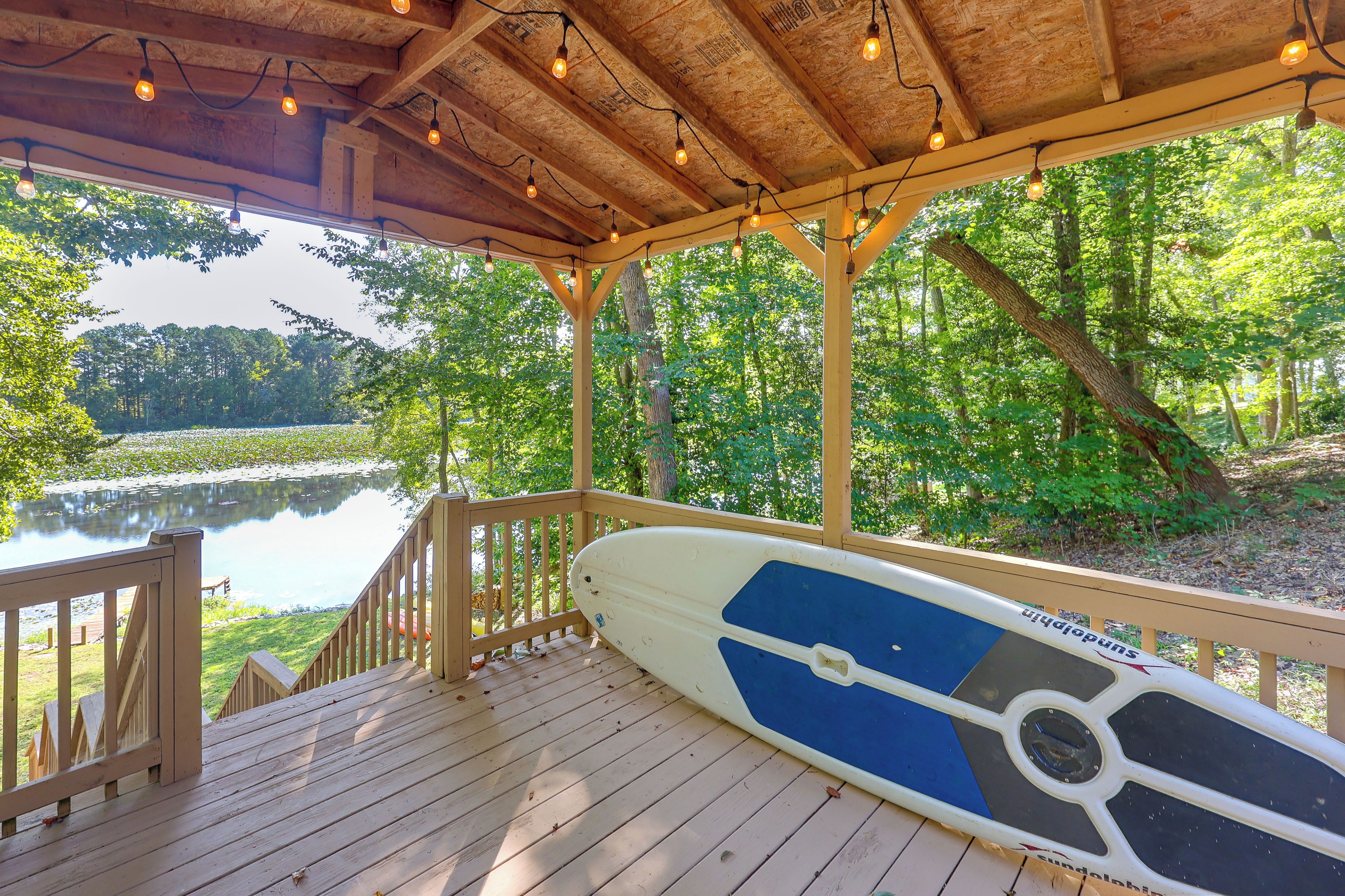 A wooden deck with a paddleboard, surrounded by trees and a body of water.