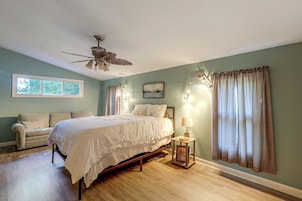 Un dormitorio con cama, mesita de noche, lámpara, ventilador de techo y ventana con cortinas.