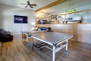Una mesa de billar en una habitación con suelo de madera, ventilador de techo y una zona de bar con taburetes y un refrigerador.