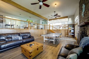 Un salón amplio con un sofá de cuero, una mesa de centro de madera y una mesa de ping pong. Una escalera conduce a un nivel superior con zona de cocina.