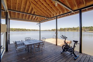 Un espacio al aire libre techado con una mesa, sillas, una bicicleta estática y vista a un lago tranquilo.
