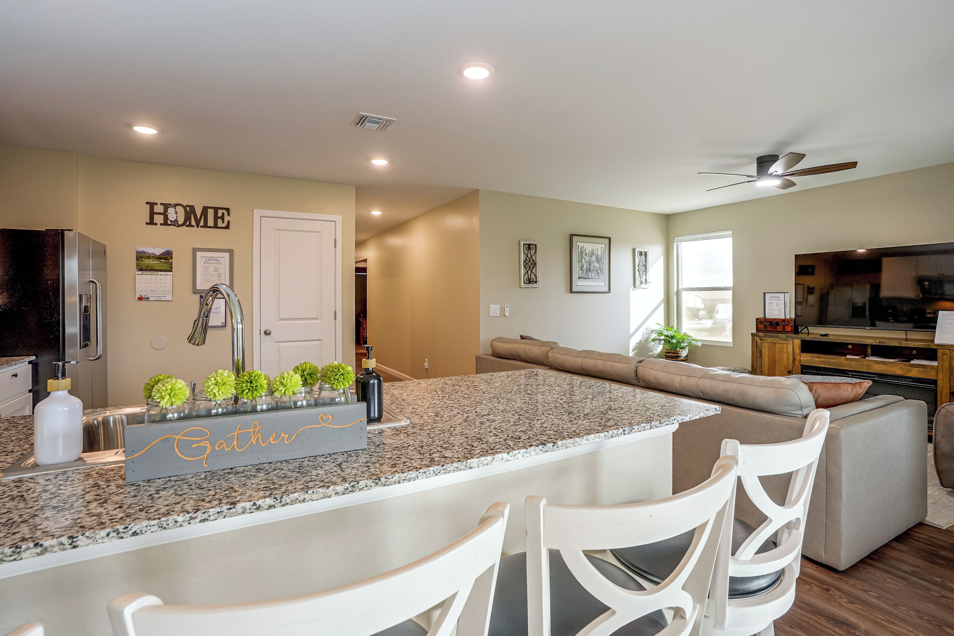 A modern kitchen with granite countertops, a built-in island, white bar stools, and a dining area with a sofa and a television.