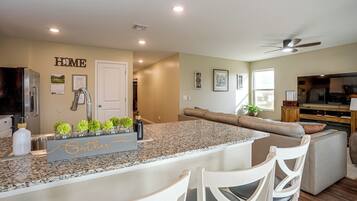 A modern kitchen with granite countertops, a built-in island, white bar stools, and a dining area with a sofa and a television.
