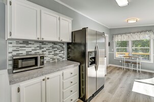 Una cocina con armarios blancos, un refrigerador de acero inoxidable, un microondas y una ventana con persianas.