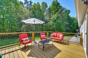 Una terraza de madera con mobiliario exterior, un paraguas a rayas y vista a un área boscosa.