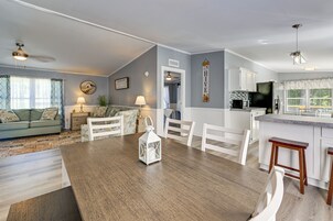 Una sala de estar moderna con una mesa de comedor de madera, sillas blancas, un sofá y una zona de cocina con armarios blancos y un refrigerador negro.