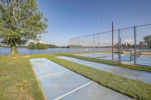 Uma quadra de tênis com cercas de arame, uma quadra de basquete e uma trilha ao lado de um lago.