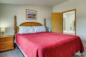 Un dormitorio con una cama de madera, una mesita de noche con lámpara, un cuadro enmarcado en la pared y una puerta de madera que da a un baño.