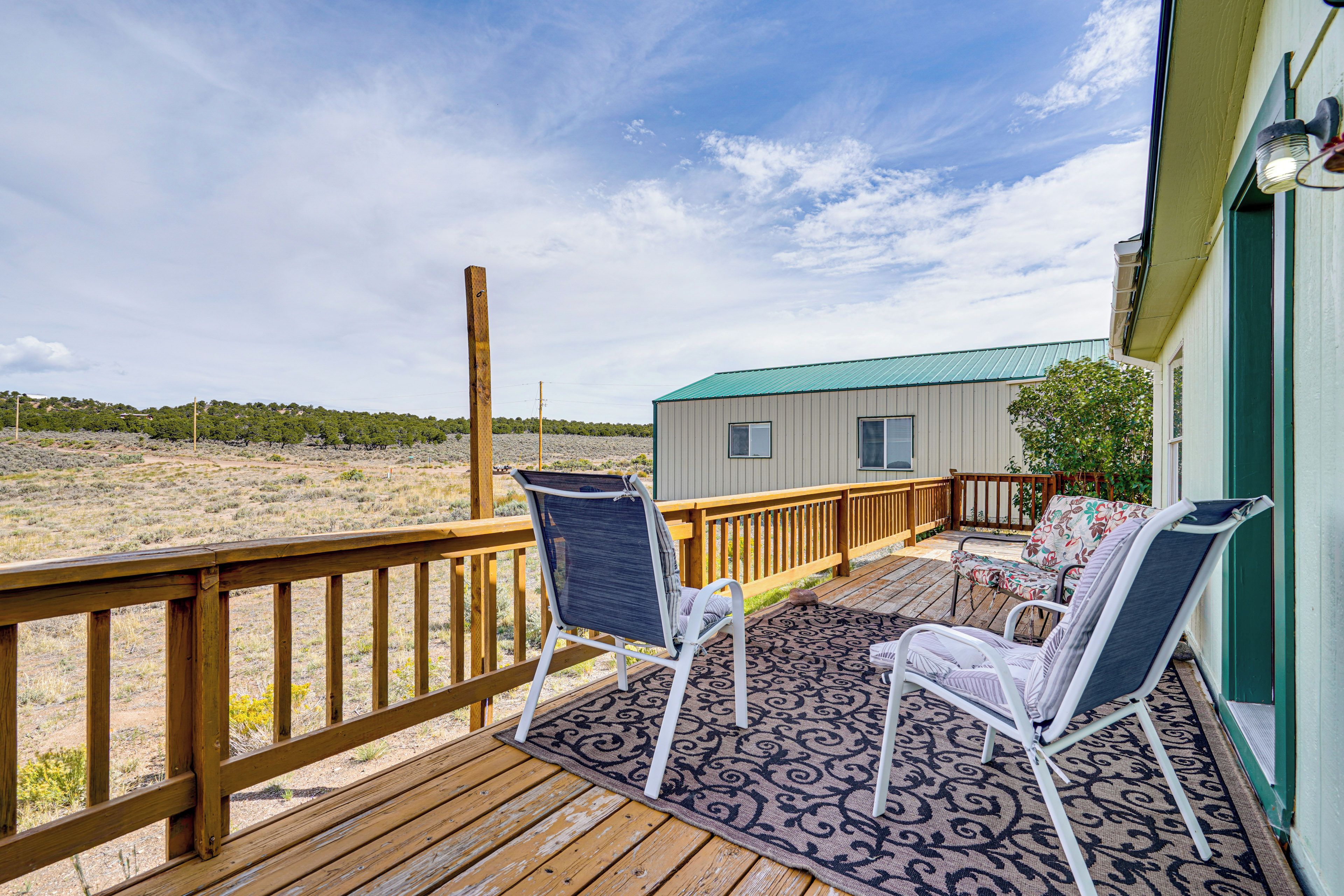 A wooden deck with two white chairs and a patterned rug, overlooking a beach and a small building with a green roof.