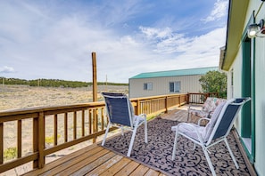 A wooden deck with two white chairs and a patterned rug, overlooking a beach and a small building with a green roof.