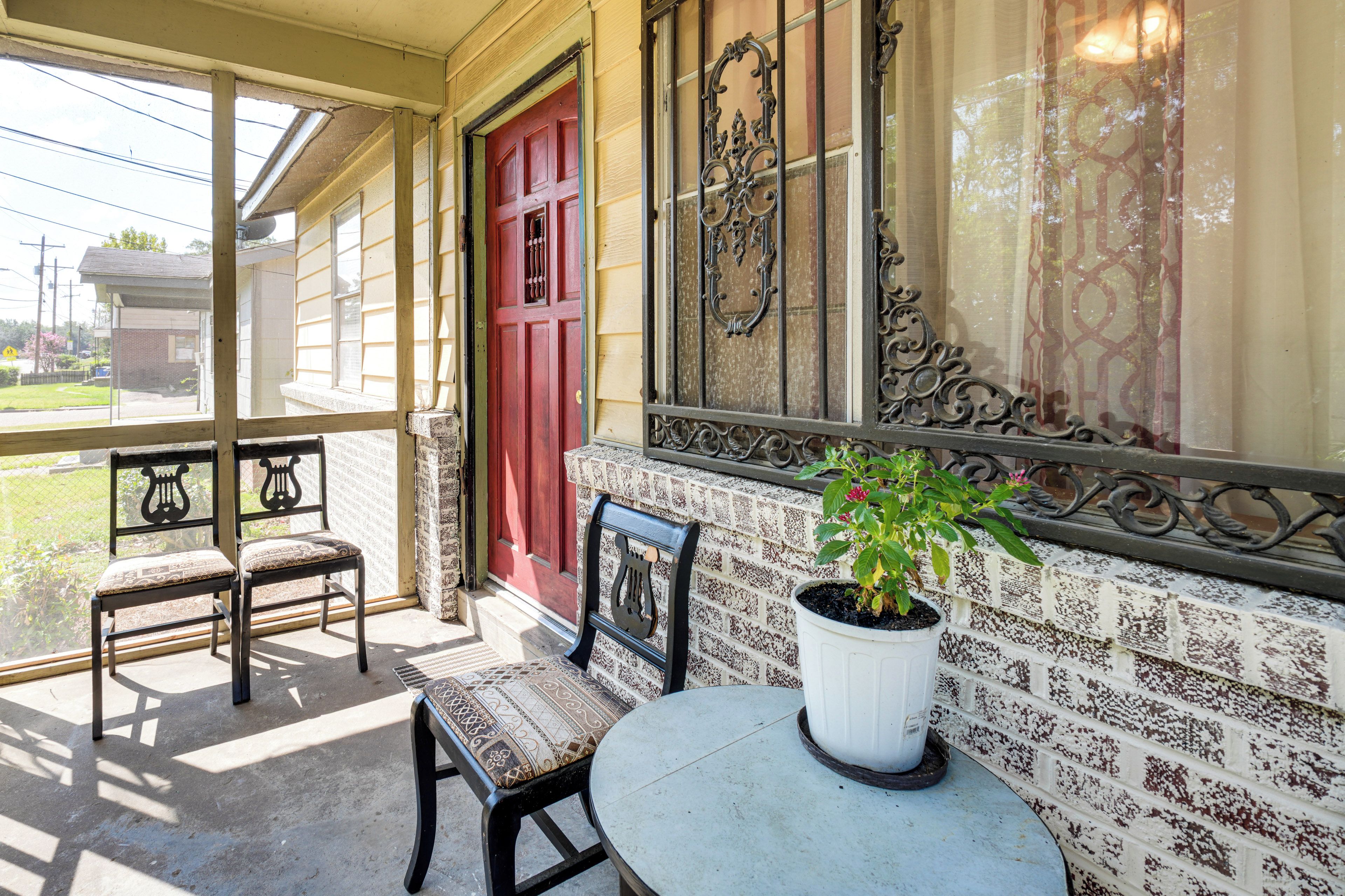 A porch with two chairs, a potted plant, and a red door.
