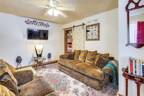A living room with a brown sofa, a glass coffee table, a patterned rug, a ceiling fan, and a bookshelf with books.