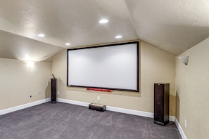 A home theater room with a large flat-screen TV mounted on the wall, carpeted floor, and ceiling-mounted lighting.