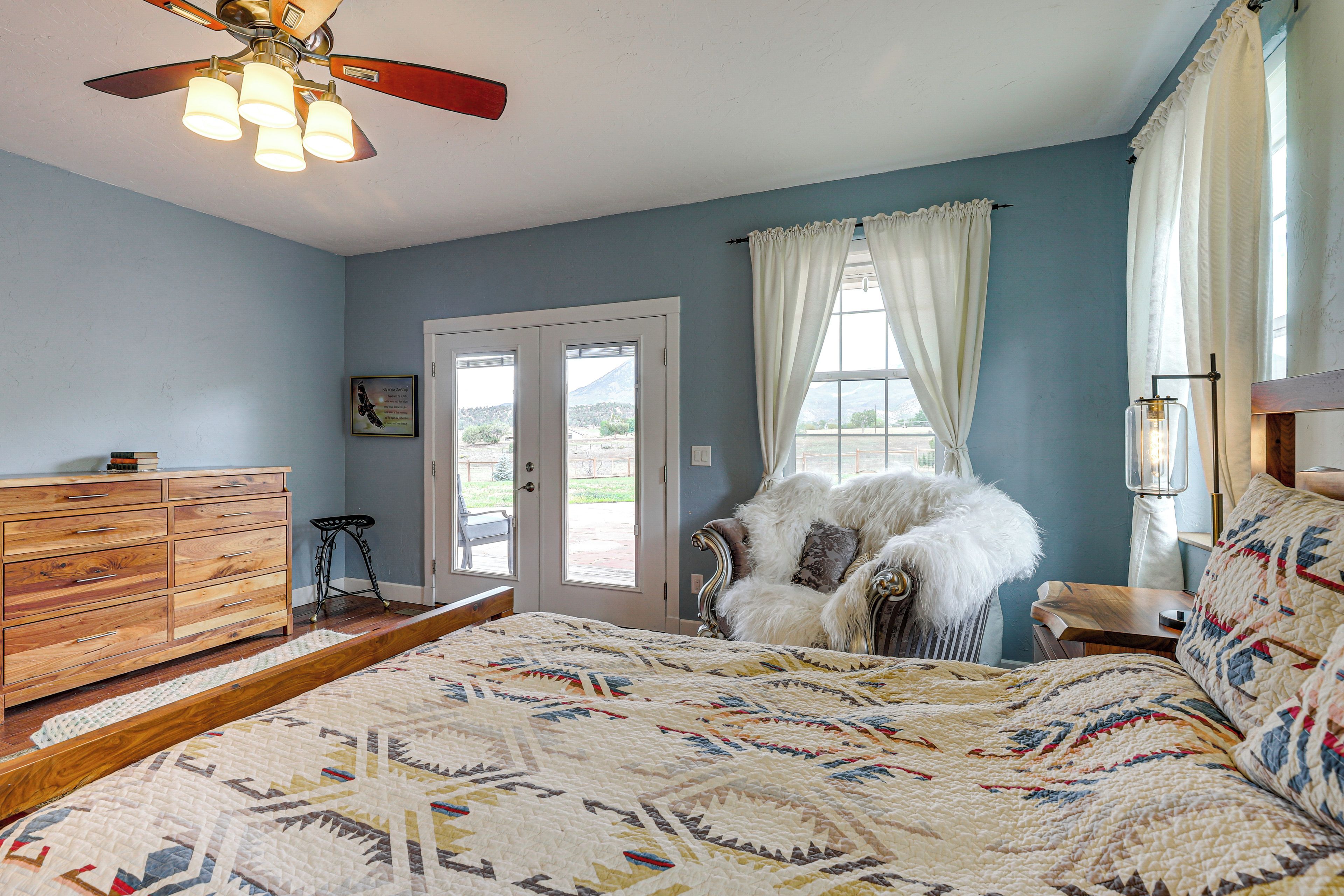 A bedroom with a large bed, a ceiling fan, a dresser, and a view of the outdoors through a door.