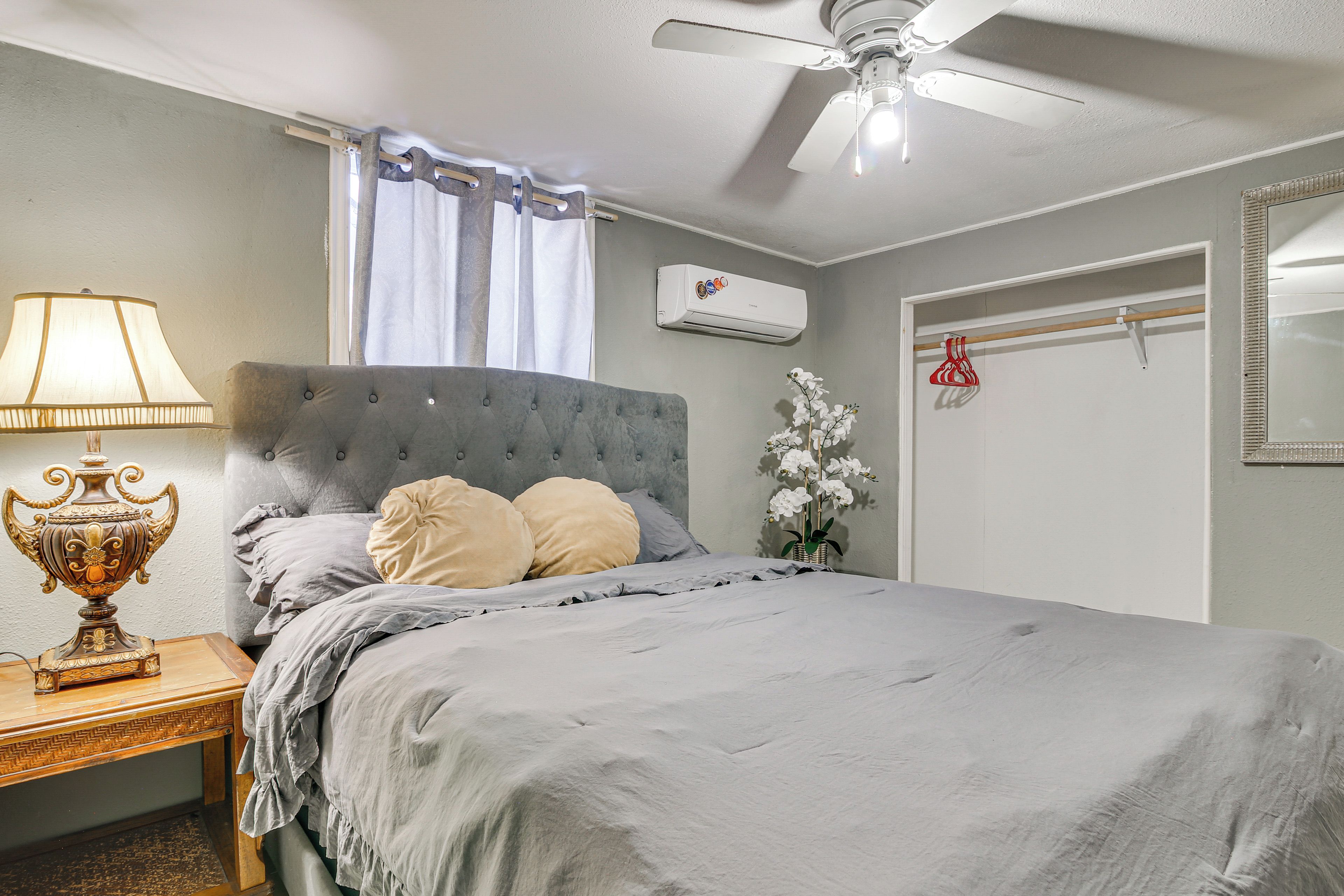 A bedroom with a bed, a nightstand, a lamp, an air conditioning unit, a window with curtains, a ceiling fan, and a mirror on the wall.