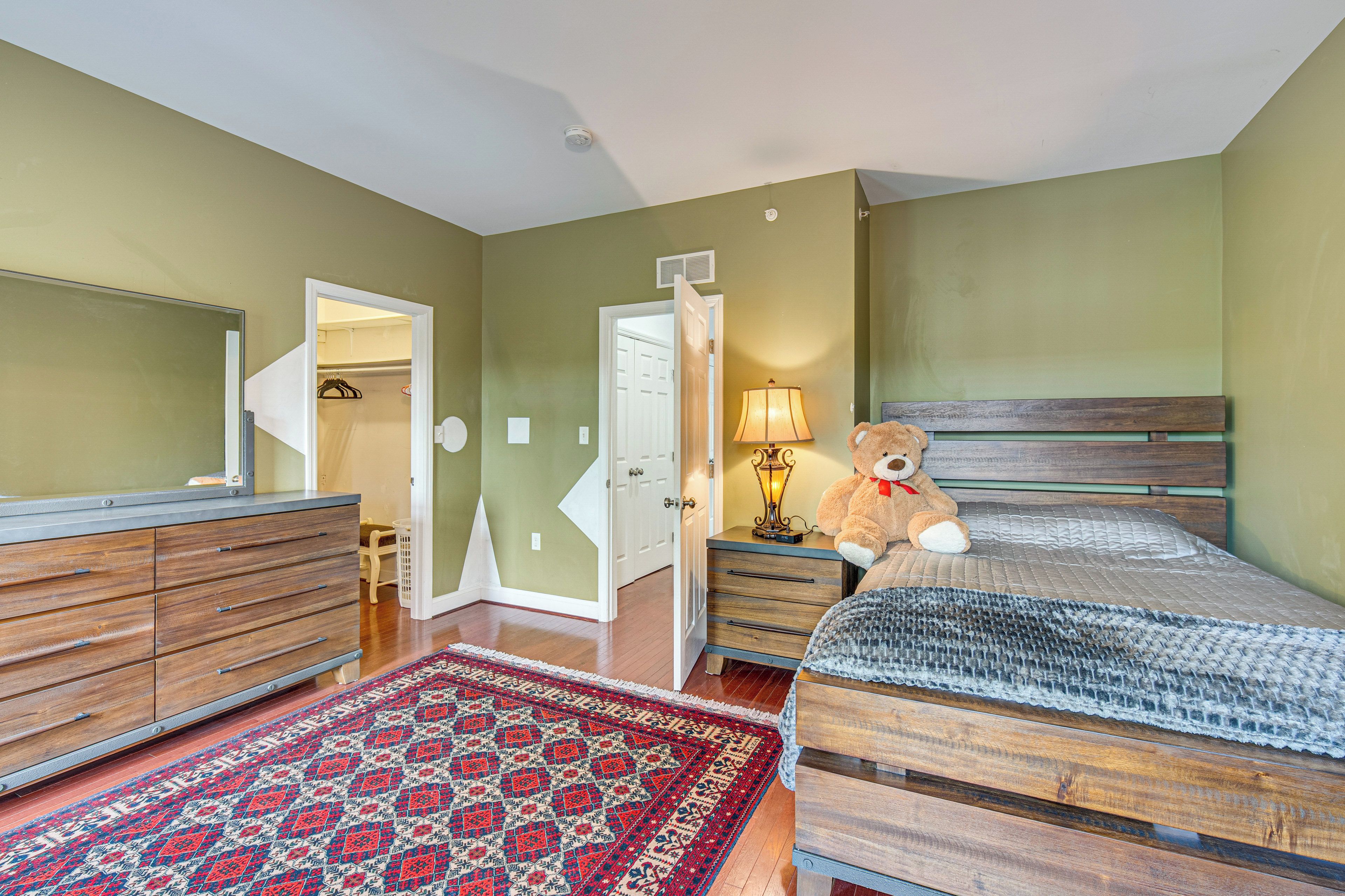 A bedroom with a bed, a dresser, a lamp, a teddy bear, and a patterned rug.