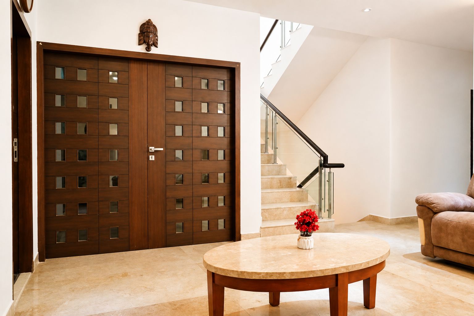 A modern interior with a wooden double door, a staircase with glass railings, a round table with a vase, and a brown sofa.