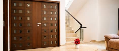 A modern interior with a wooden double door, a staircase with glass railings, a round table with a vase, and a brown sofa.