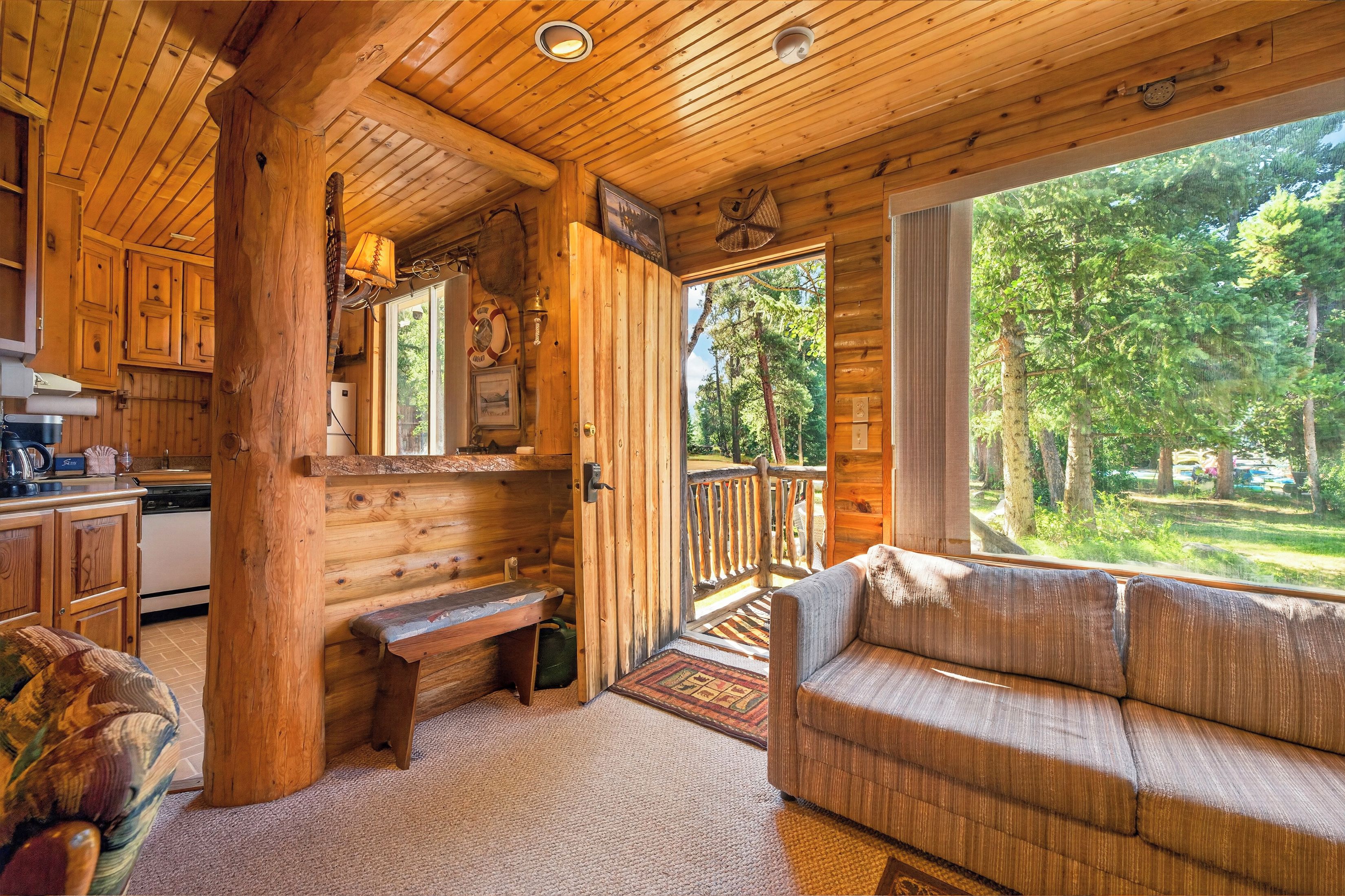 A cabin interior with a wooden ceiling, a sofa, a kitchen area, and a view of the forest.