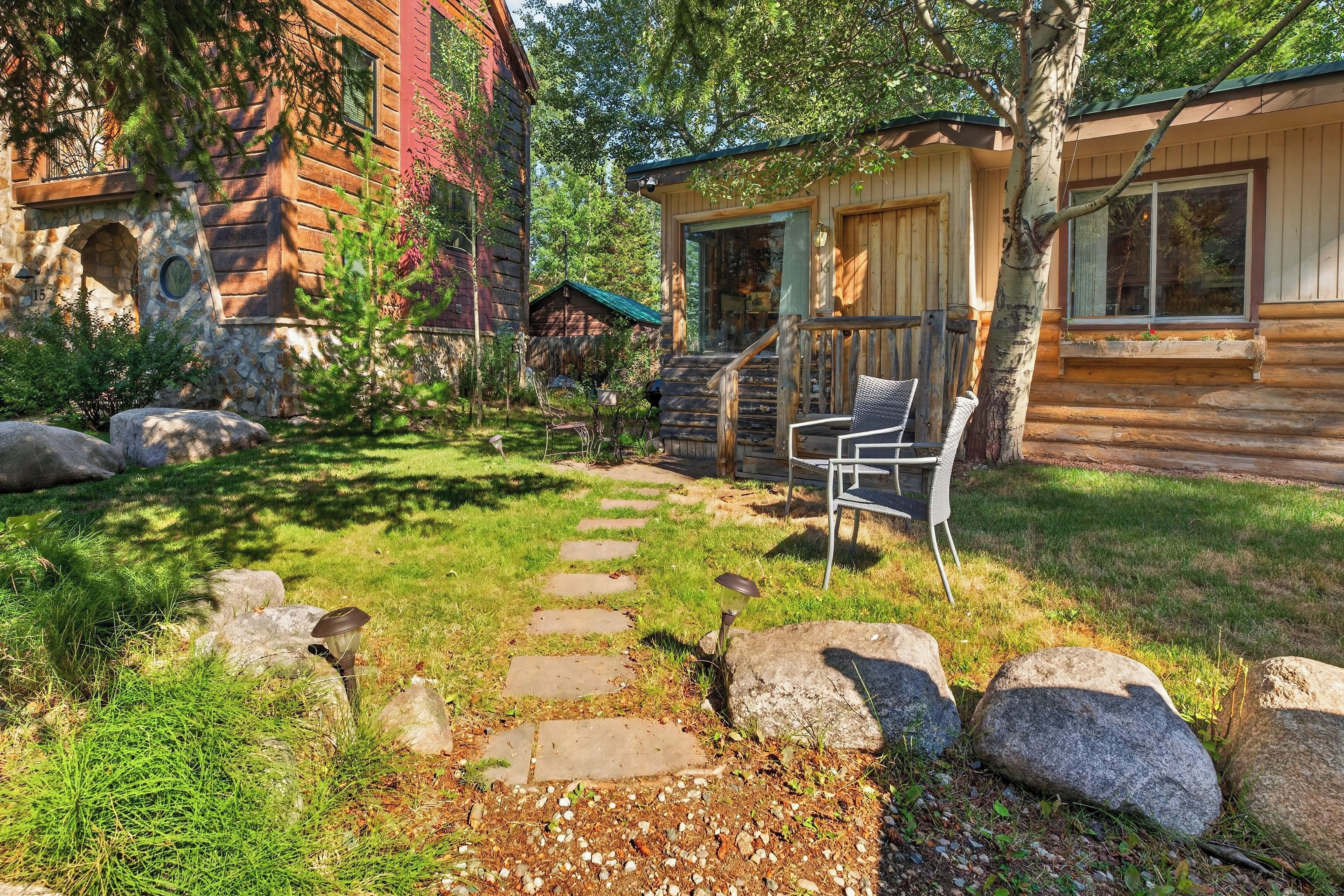 A rustic cabin with a stone pathway leading to a wooden deck, surrounded by rocks and greenery.