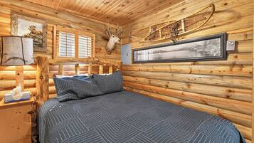 Ein Schlafzimmer in einer Blockhütte mit einem Bett, einem aufgehängten Hirschkopf, einem gerahmten Bild und einem Fenster mit Jalousien.