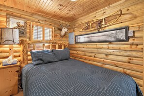 Ein Schlafzimmer in einer Blockhütte mit einem Bett, einem aufgehängten Hirschkopf, einem gerahmten Bild und einem Fenster mit Jalousien.
