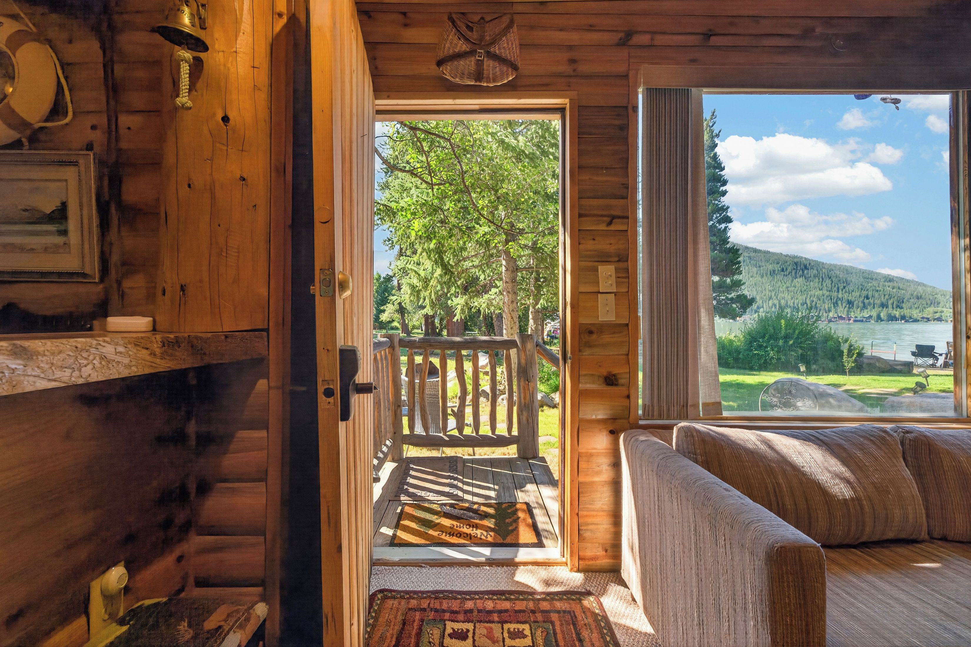 A cabin interior with a wooden door leading to a deck, a sofa, and a view of a lake and forest.