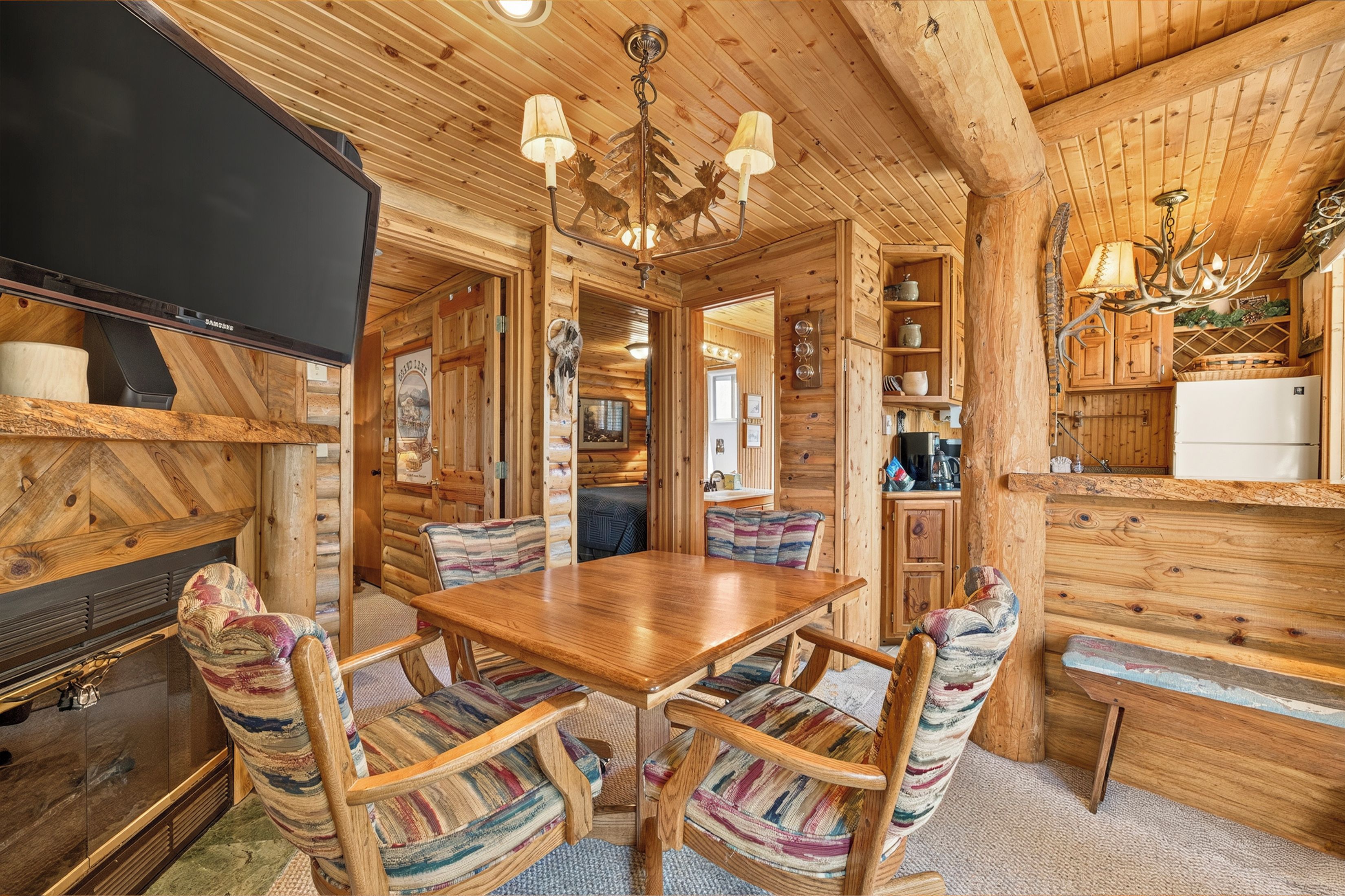 A log cabin interior with a wooden dining table, chairs, a television, and a kitchen area.