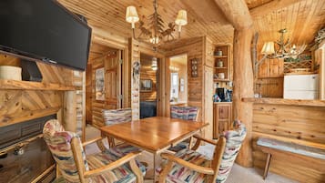 A log cabin interior with a wooden dining table, chairs, a television, and a kitchen area.