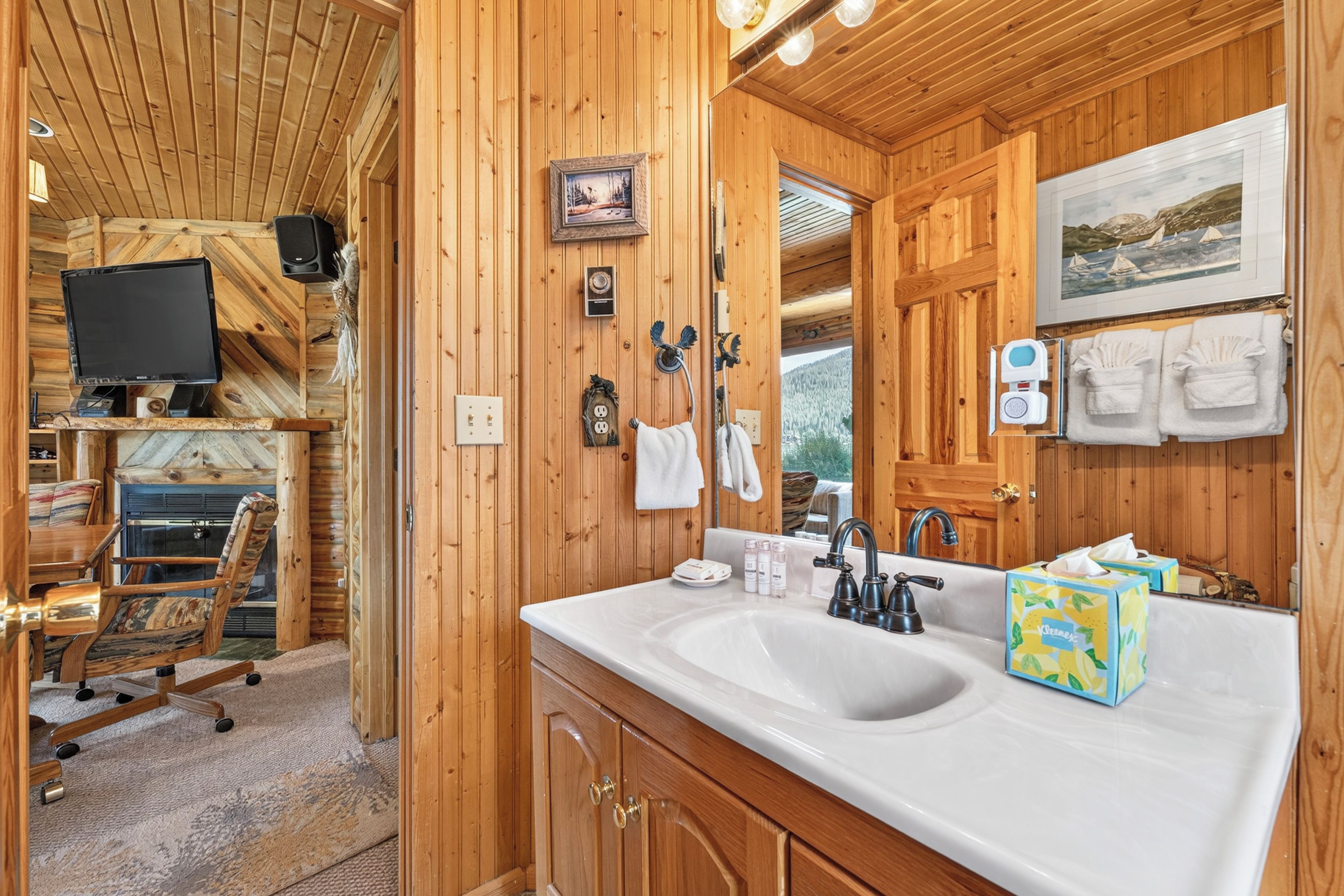 Ein Interieur einer Holzhütte mit einem großen Spiegel mit Holzrahmen, einem weißen Waschbecken und einem an der Wand befestigten Fernseher.