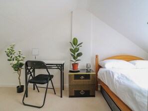 Ein Schlafzimmer mit einem Bett, einem Schreibtisch mit Stuhl, einer Topfpflanze und einem Nachttisch mit einem Glas.