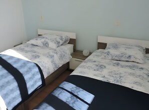 A room with two beds, each with a patterned bedspread and pillows, a bedside table, and a wall-mounted light fixture.