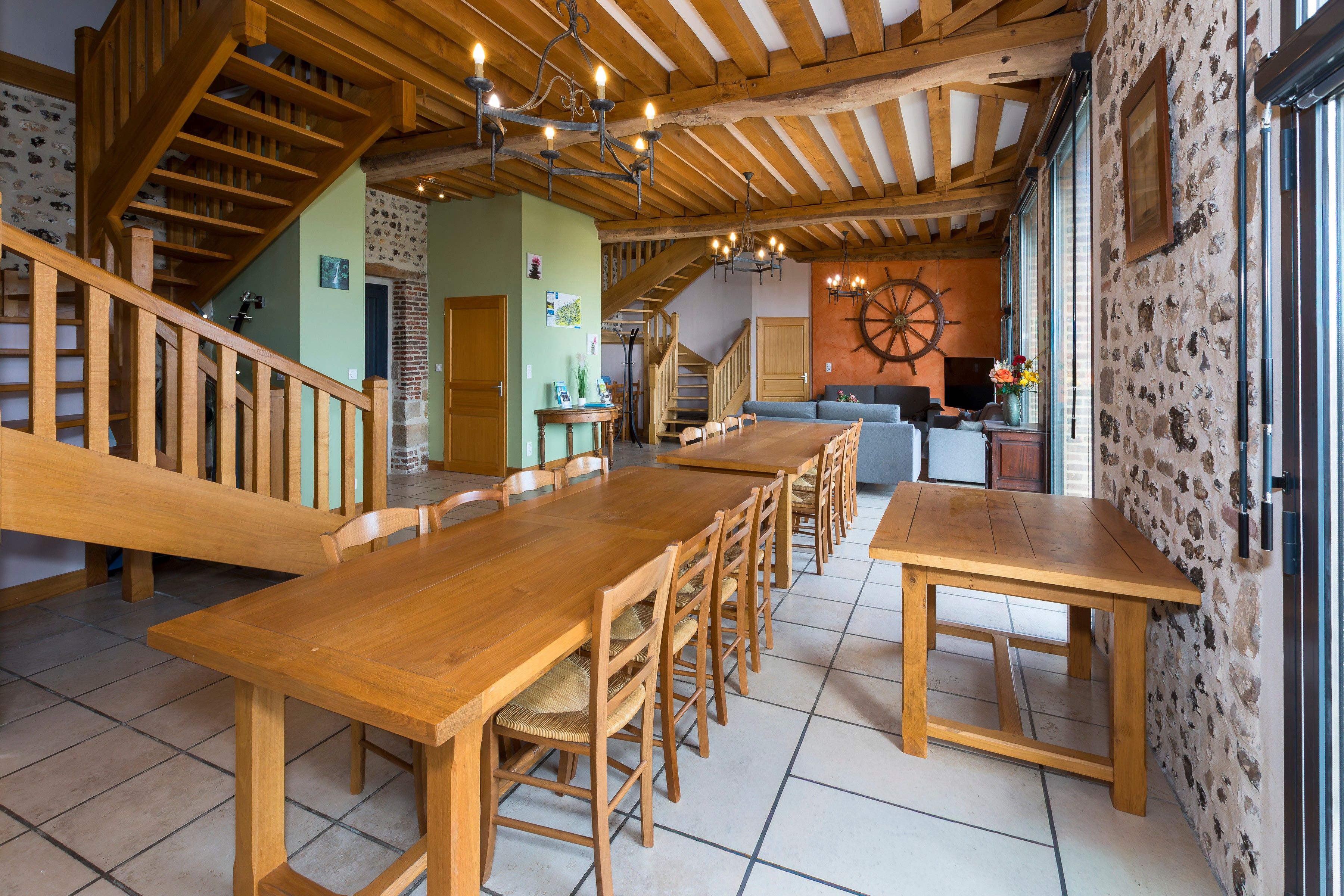 A spacious dining area with wooden tables and chairs, a staircase, and a rustic stone wall.
