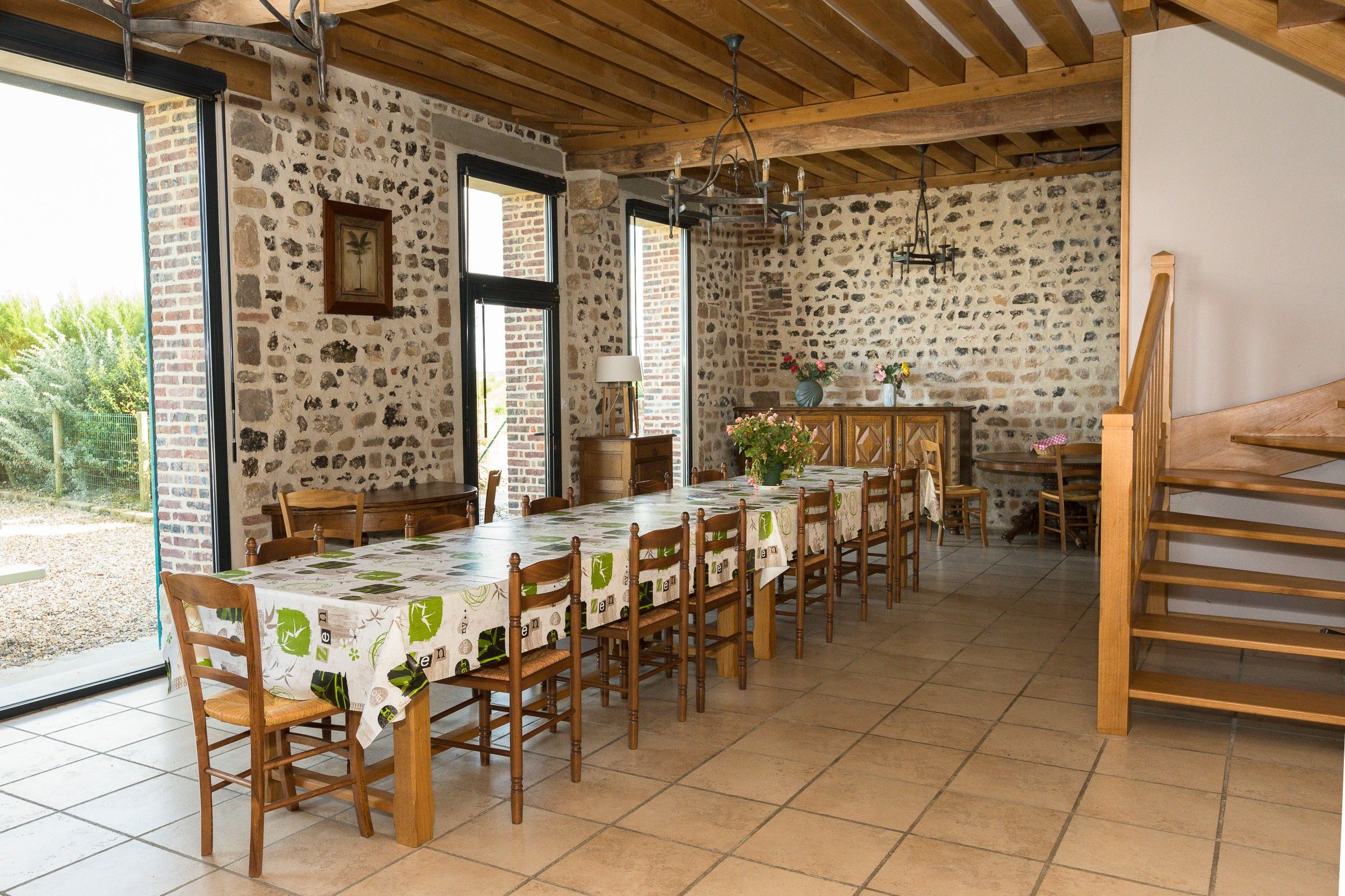 Un espace repas avec une longue table dressée pour accueillir plusieurs personnes, des chaises en bois, un lustre et un mur en pierre.