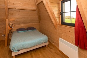 A single bed with a blue mattress and two pillows in a wooden room with a sloped ceiling and a window with red curtains. - Cottage 'Le Four À Pain' with Private Terrace and Wi-Fi (Saint-Aubin-sur-Mer)