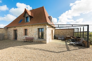 A stone house with a tiled roof, a covered patio with outdoor furniture, and a gravel driveway. - Cottage 'Le Four À Pain' with Private Terrace and Wi-Fi (Saint-Aubin-sur-Mer)