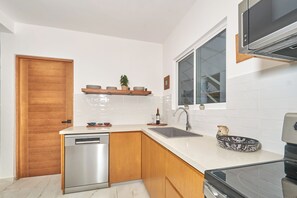 Cocina compacta con lavavajillas, pileta y estufa. Se observa una puerta de madera, una ventana y un estante con vajilla.
