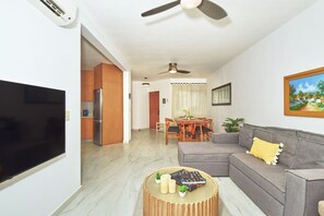 Una sala moderna con un televisor de pantalla plana, un sofá gris, una mesa de comedor de madera y un ventilador de techo.