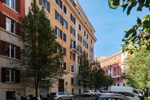 Straßenansicht mit mehrstöckigen Gebäuden, parkenden Autos und Bäumen.