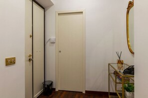 A hallway with a closed door, a trash bin, a light switch, and a small table with a plant and a decorative item. - A roomy and comfy 100 sqm apartment, ideal for four people, on the second floor of a building (with elevator; though NOT suitable for people with disabilities due to several steps at the entrance). It is situated in the elegant and quiet Della Vittoria d (Rome)