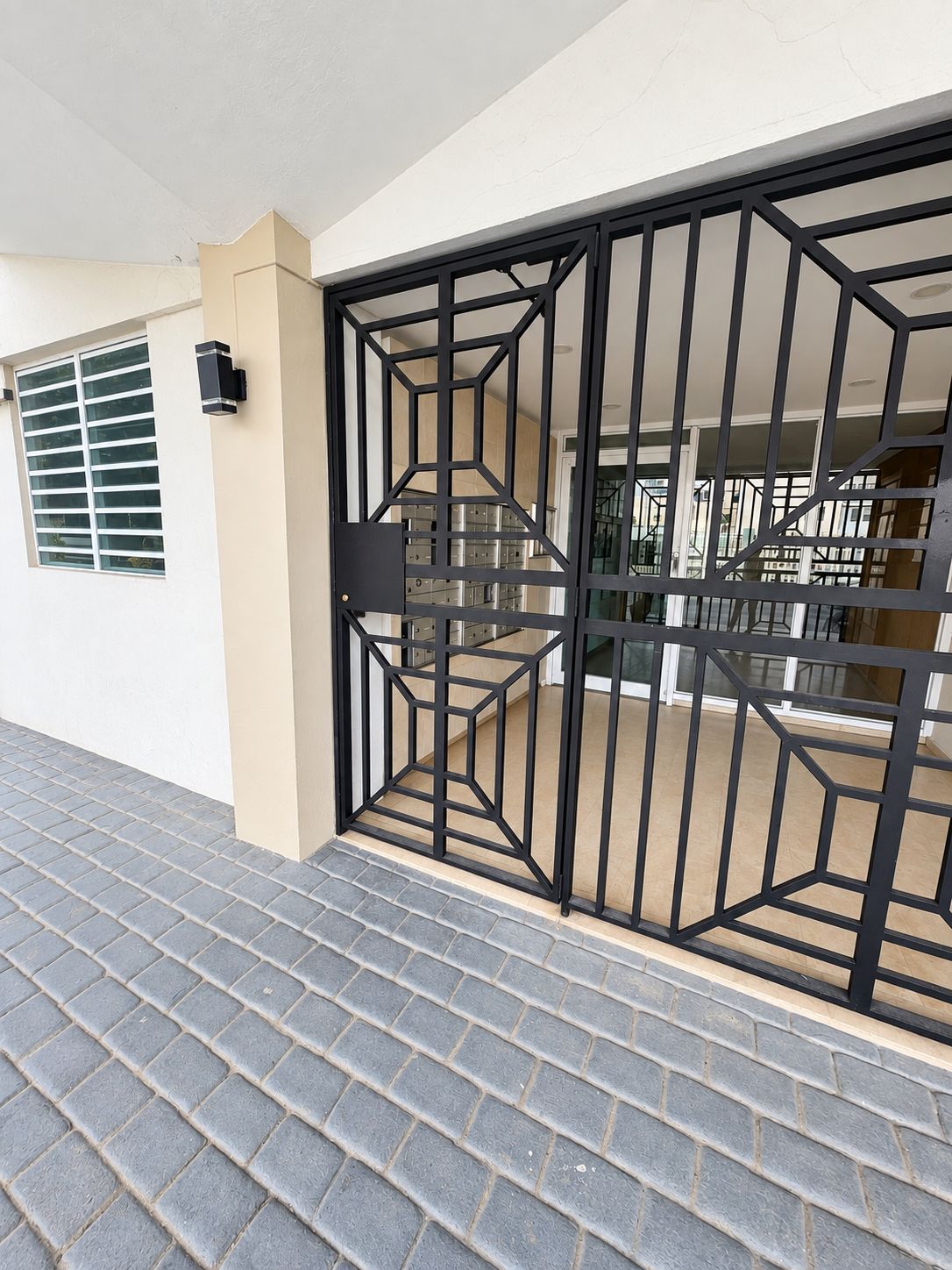 A gated entrance with a geometric metal design, a window with horizontal blinds, and a paved walkway.