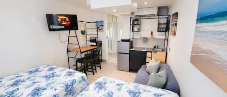 Habitación compacta con dos camas, una kitchenette, un pequeño comedor y un televisor fijado a la pared.