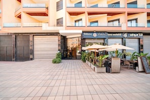 Een meerlaags gebouw met een commerciële ruimte op de begane grond, voorzien van buiten zitplaatsen en potplanten.