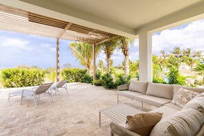 Een overdekt terras met een bank en stoelen, omringd door groen en palmbomen.