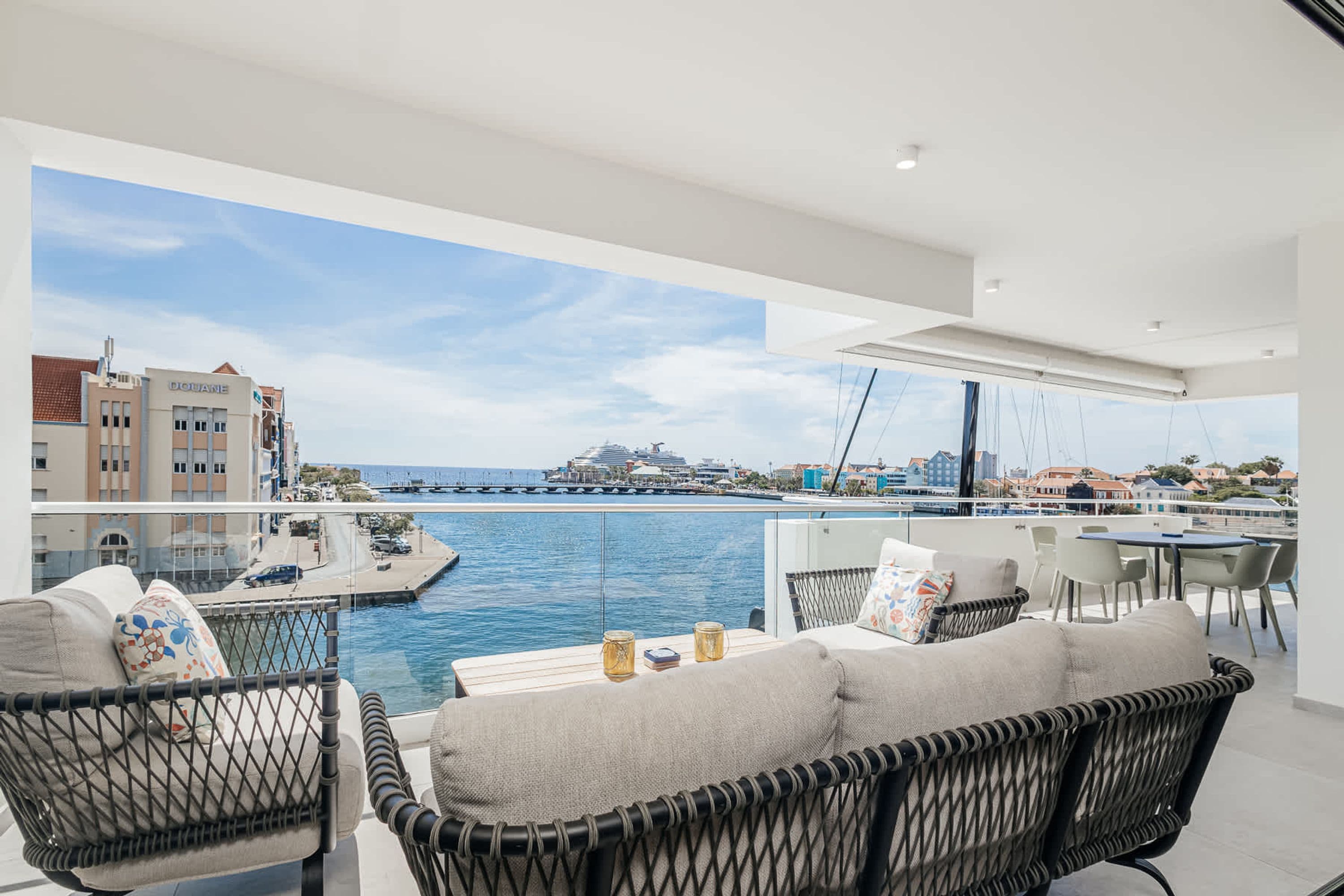 A modern balcony with wicker furniture, a view of the sea, and a marina in the background.