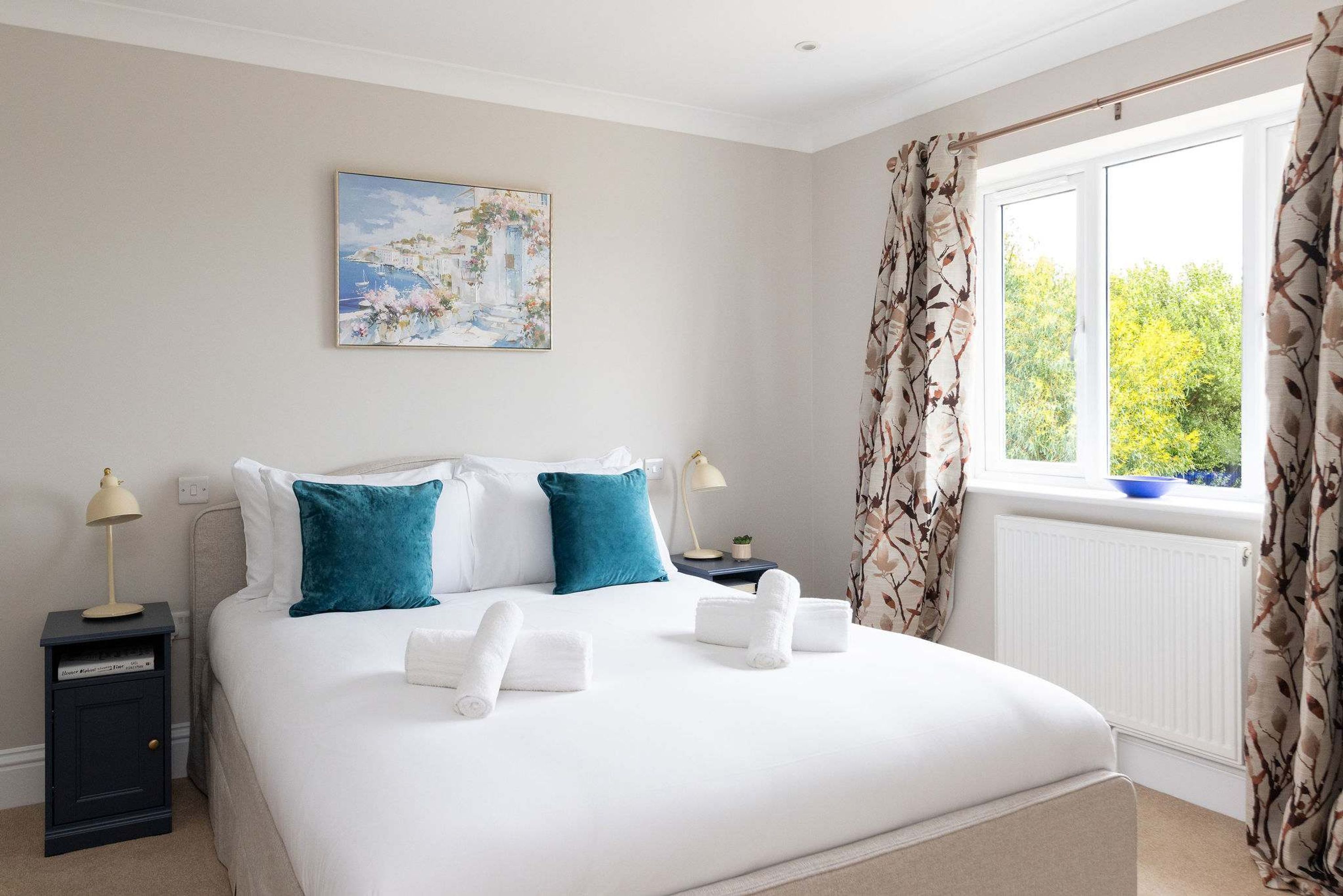 A bedroom with a bed, pillows, a nightstand, a lamp, a painting, and a window with curtains.