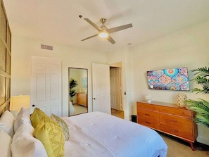 A bedroom with a bed, a dresser, a ceiling fan, a mirror, and a wall-mounted artwork. - Casa Arenas Modern Desert Retreat in Old Town LQ (La Quinta)
