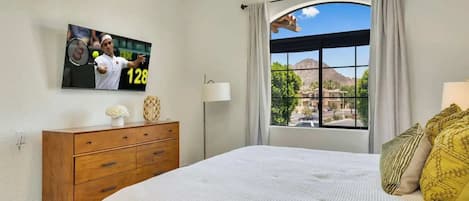 A bedroom with a bed, a dresser, a TV mounted on the wall, and a window with a view of the outdoors.