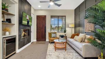 A modern living room with a fireplace, a television, a ceiling fan, and a large window with blinds.